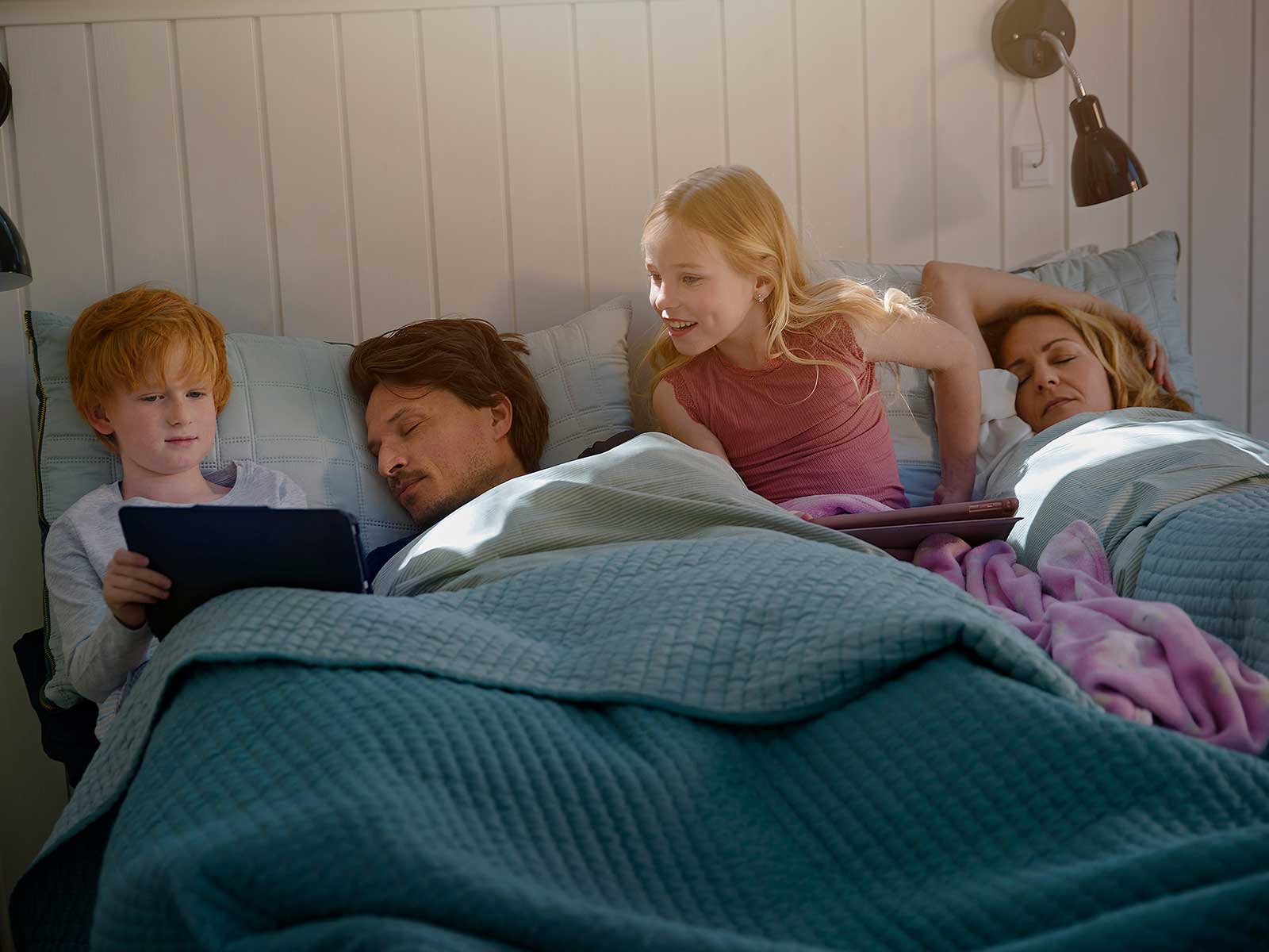 Familie ligger i sengen. Børnene med tablets.